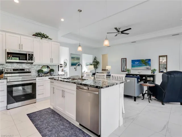 Kitchen With White Shaker Cabinetry, Kitchen Island With Double Stainless-Steel Sink, Pendant Lighting, Glass Tile Backsplash, & Glass Tile Backsplash, & Living Room With Tray Ceiling and Ceiling Fan