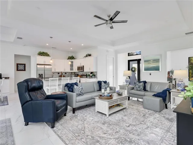 Living Room With Tray Ceiling & Ceiling Fan, Kitchen With White Shaker Cabinetry, Island, Glass Tile Backsplash,  Stainless-Steel Appliances, and Quartz Counters