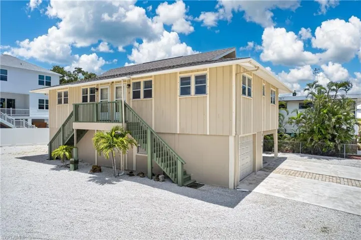 Raised coastal-style residence featuring vertical siding, dual exterior staircases, ground-level garage access, a gravel lot, and a paver driveway