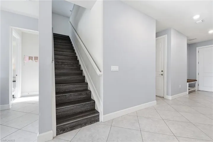 Wood-finish staircase with white handrail, light gray wall tones, white trim, and light-toned tile flooring throughout
