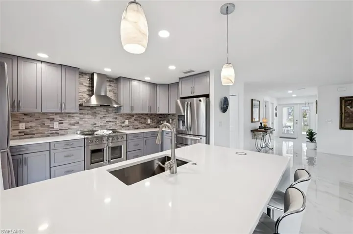 Kitchen featuring gray cabinetry, wall chimney range hood, stainless steel appliances, pendant lighting, and light stone counters