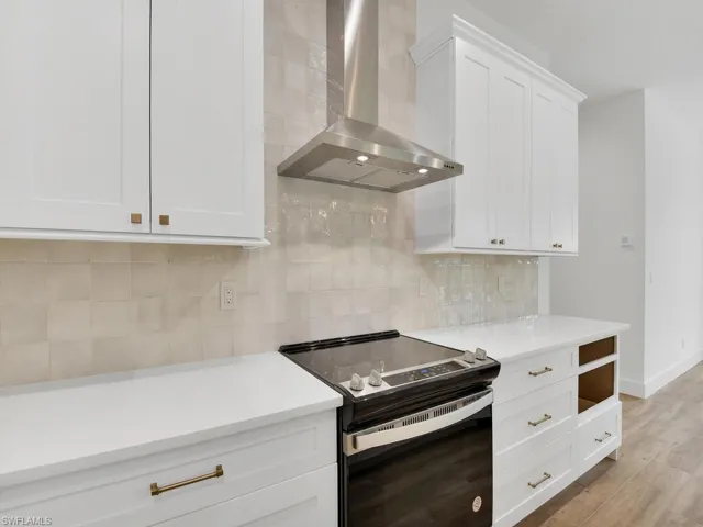 Kitchen featuring electric range, wall chimney range hood, white cabinets, decorative backsplash, and light stone counters