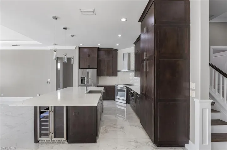 Kitchen featuring decorative light fixtures, appliances with stainless steel finishes, a large island with sink, light stone counters, and wall chimney range hood