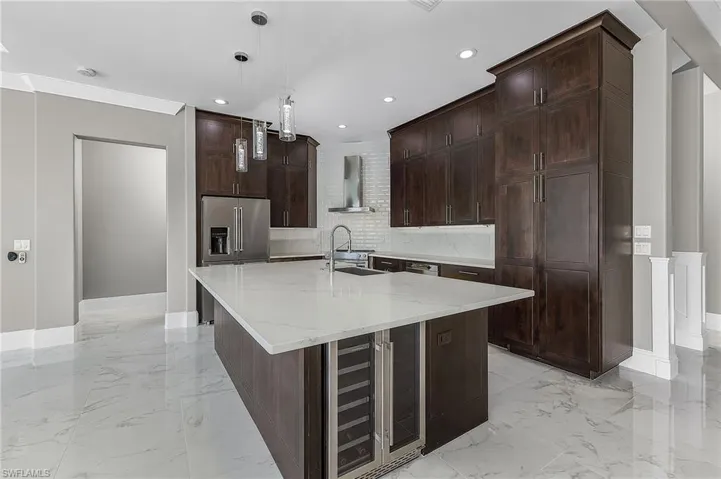 Kitchen featuring wine cooler, an island with sink, light stone countertops, hanging light fixtures, and wall chimney range hood