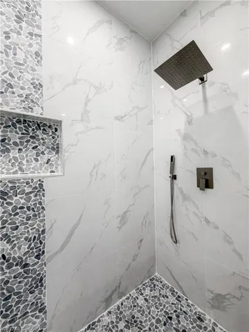 Shower enclosure featuring large format white marble-look wall tiles, pebble-tiled accent wall and flooring, square rain shower head, and built-in shower niche