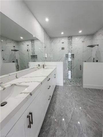 Spacious bathroom featuring a dual vanity with a white countertop and integrated sinks, extensive wall-to-wall mirror, two glass-enclosed shower stalls with rain showerheads, and large-format grey-toned floor tiling