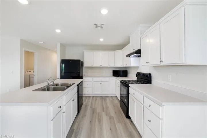 Kitchen featuring washer and dryer, black appliances, white cabinets, light wood-type flooring, and an island with sink