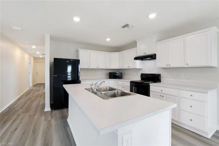 Kitchen with black appliances, light wood finished floors, white cabinetry, an island with sink, and recessed lighting