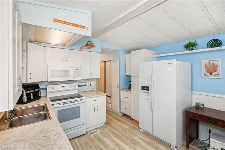 White shaker-style cabinetry, double basin stainless steel sink, wood-finish flooring, white appliances, and a built-in microwave