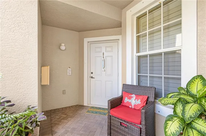 Covered entryway featuring a white paneled door, large grid window, stucco exterior, wall-mounted light fixture, and brick paver flooring