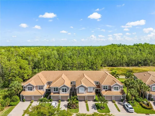 Townhome complex featuring tan tile roofs, gray siding, and attached garages, with a dense tree line backdrop