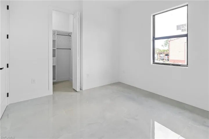 Spacious room featuring light-toned flooring, crisp white walls, a large window with dark trim, a built-in closet with shelving, and a door with dark hardware