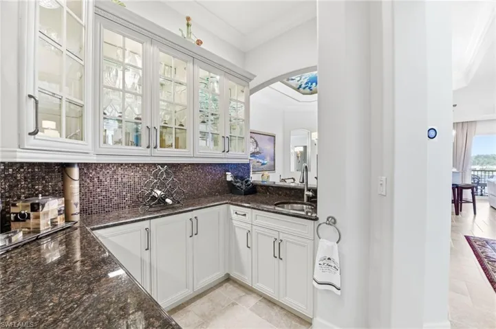 Bar with ornamental molding, dark stone countertops, white cabinetry, glass fronted cabinets, and arched walkways