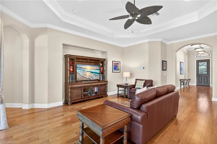 Open living area with tray ceiling, crown molding, hardwood floors, ceiling fan, and defined foyer entry.