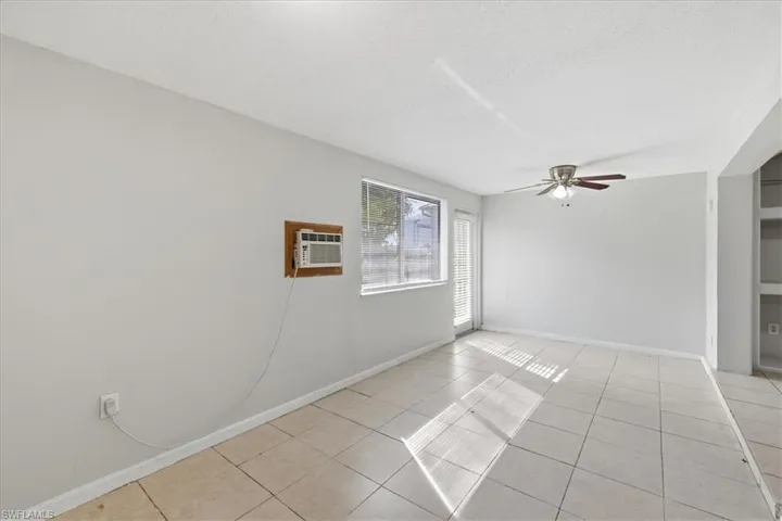Bright living space featuring light-toned tile flooring, a window-mounted air conditioning unit, a ceiling fan with light fixture, a window with horizontal blinds, and a built-in shelving unit