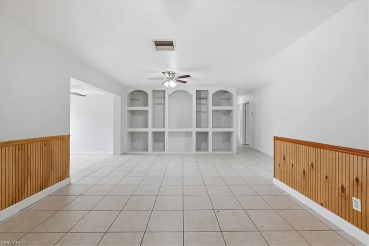 Open room featuring light-toned tile flooring, built-in shelving with arched accents, wood paneling on lower walls, and a ceiling fan with integrated lighting