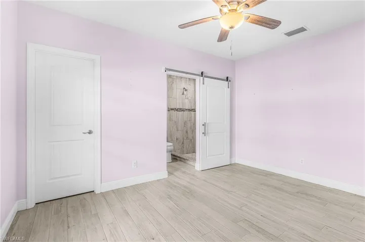 Unfurnished bedroom featuring a barn door, light wood finished floors, ensuite bath, and ceiling fan