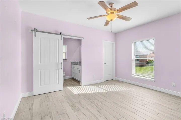 Unfurnished bedroom featuring a barn door, light wood-type flooring, ensuite bath, and a ceiling fan