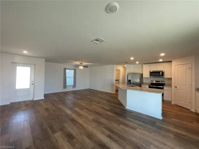 Kitchen with dark wood-type flooring, white cabinets, light stone countertops, an island with sink, and stainless steel appliances