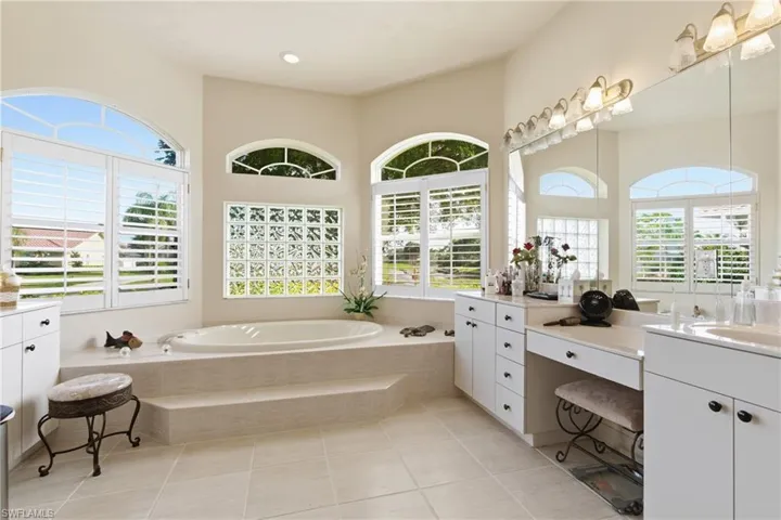 Full bathroom featuring duel sink vanities, jacuzzi tub, light tile patterned flooring, plantation shutters, and healthy amount of natural light