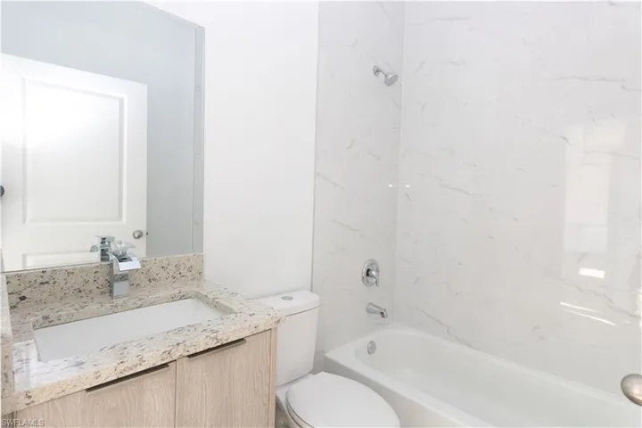 Bathroom featuring a light wood-finish vanity, a speckled stone countertop, a white rectangular sink, a polished chrome faucet, and a white tiled shower surround with a built-in tub