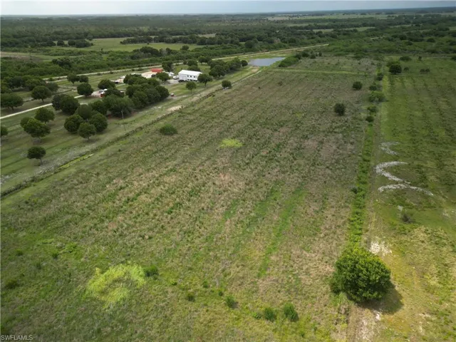 Expansive land featuring a white building, multiple mature trees, a visible pond, and a large open field