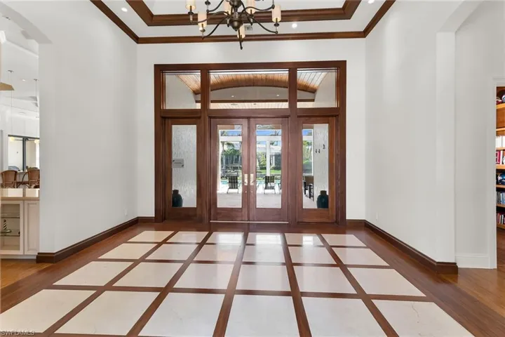 Grand entry foyer featuring a coffered ceiling, wood-finish flooring with light-toned inset tiles, dark wood trim, and double glass-paneled doors with transom windows