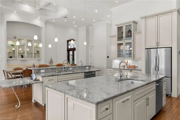 Gourmet kitchen featuring multiple granite-top islands, stainless steel appliances, extensive cabinetry with glass display fronts, recessed lighting, and hardwood flooring