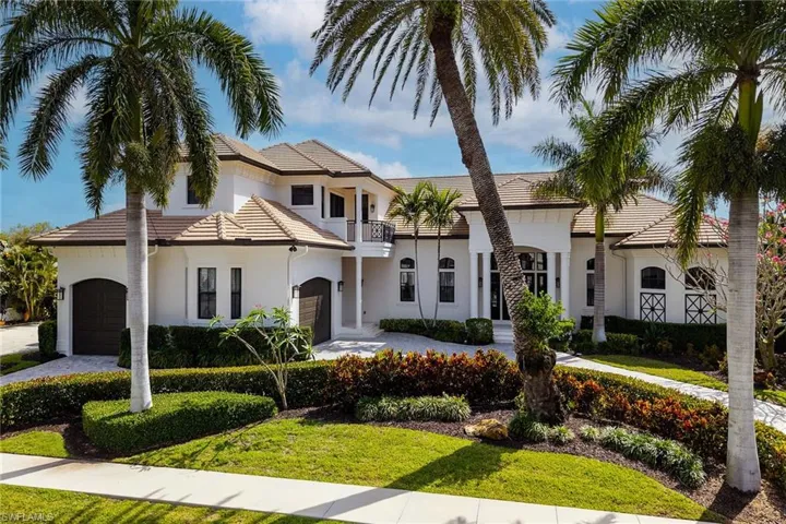 Elegant white stucco residence featuring a tile roof, dual arched garage doors, a columned entryway, mature palm trees, and manicured landscaping
