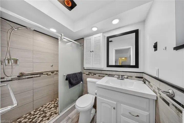 The bathroom features a tiled shower with a sliding glass door and pebble-style floor, a white vanity with an integrated sink, a framed mirror, and recessed lighting