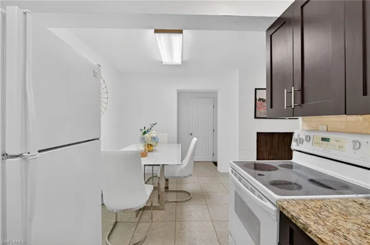 Kitchen featuring white appliances, dark wood finish cabinetry, light tile patterned flooring, and light stone counters