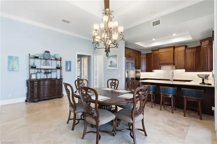 Tile flooring, a high ceiling with crown molding, and a chandelier illuminate a spacious kitchen with wood cabinetry, a built-in oven, and a white tile backsplash - Virtually Edited Image