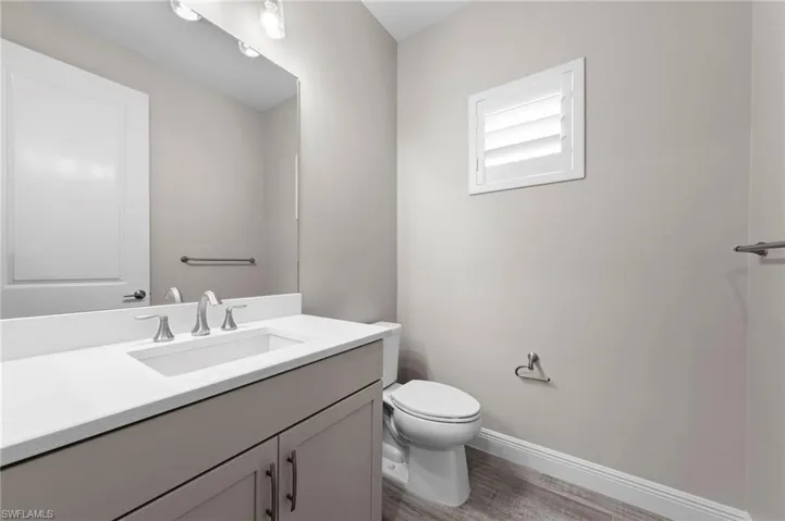 Bathroom featuring a single-sink vanity with a light countertop, wood-finish flooring, a white toilet, and a window with white louvered shutters