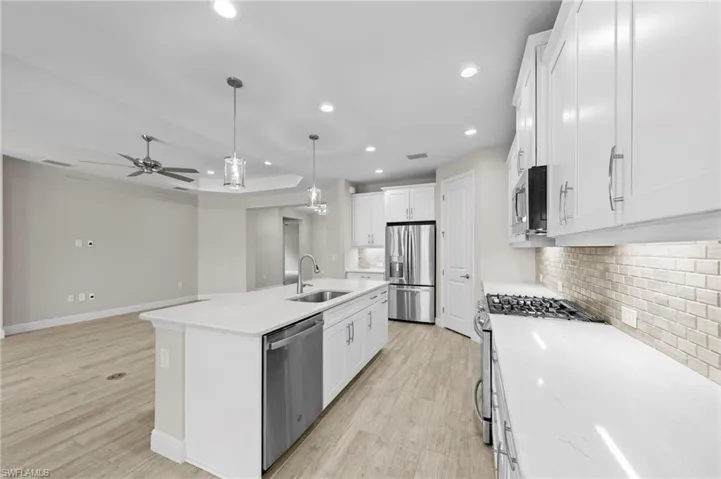 Open-concept kitchen featuring an expansive island with a sink and pendant lighting, white shaker cabinetry, stainless steel appliances, and wood-finish flooring