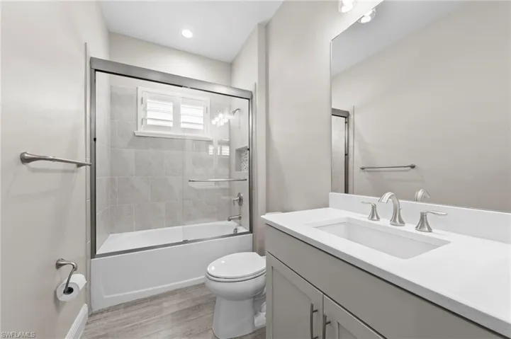 Bathroom featuring a white vanity with an integrated sink, brushed nickel fixtures, a tub with a glass shower enclosure, light gray tiled walls, and light wood-finish flooring