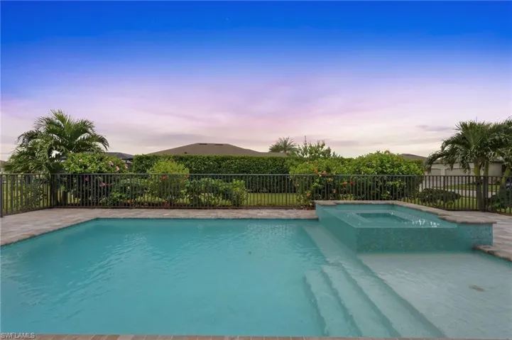 Outdoor swimming pool with integrated spa, tile pool steps, and paver decking, surrounded by a metal fence and mature landscaping