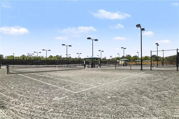 Multiple tennis courts featuring asphalt surfacing, perimeter fencing, overhead lighting, and shaded seating areas