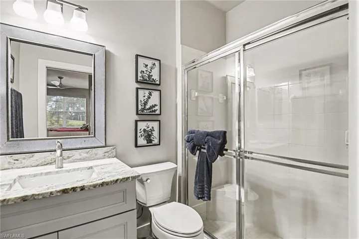 Bathroom featuring a single-sink vanity with a stone countertop, a framed mirror, a toilet, and a glass-enclosed shower with white tiled walls and a built-in bench