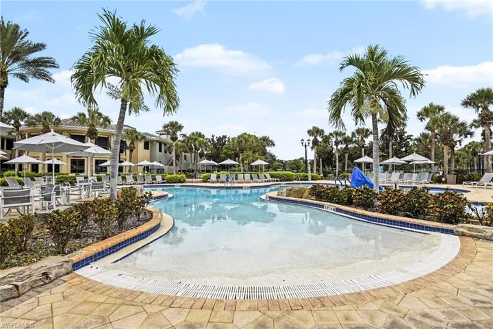 Resort-style pool area featuring a freeform swimming pool with a gradual entry, patterned paver decking, surrounding lounge seating with umbrellas, mature palm trees, and lush landscaping