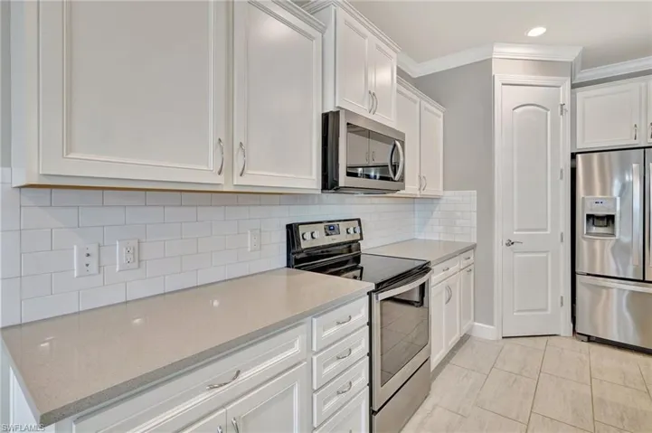 Kitchen featuring light stone countertops, appliances with stainless steel finishes, white cabinetry, decorative backsplash, and ornamental molding