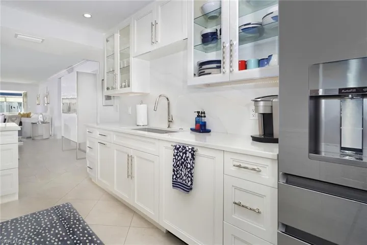 Kitchen with stainless steel fridge, glass insert cabinets, white cabinets, light tile patterned floors, and light stone counters