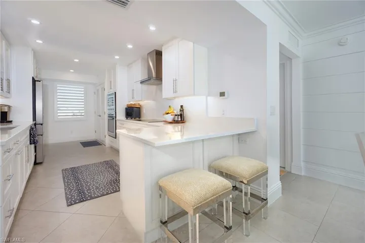 Kitchen featuring white cabinets, light stone countertops, a peninsula, wall chimney range hood, and a kitchen breakfast bar