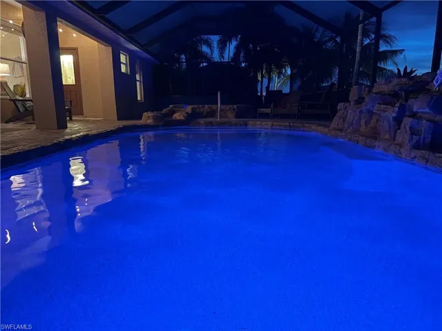 Screened swimming pool with blue interior lighting, rock waterfall feature, and surrounding stone coping