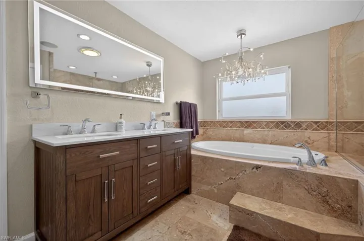 Full bath featuring double vanity, a garden tub, a textured wall, hanging lights, and light stone finish flooring