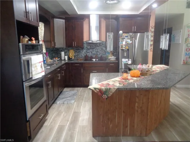 Kitchen with wall chimney range hood, stainless steel appliances, granite counters, light wood finished floors, and dark brown cabinets