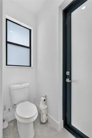 Compact bathroom featuring a white toilet, frosted-pane window with black framing, sleek black door frame, white baseboards, and grey tile flooring