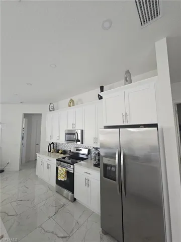 Kitchen featuring appliances with stainless steel finishes, light marble finish flooring, tasteful backsplash, white cabinetry, and light countertops
