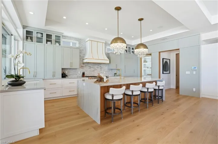 Kitchen with glass fronted cabinets, light wood-type flooring, light stone countertops, a large island with sink, and a tray ceiling - Virtually Edited Image