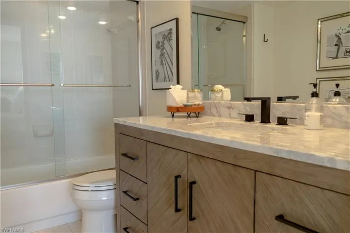 The bathroom features a vanity with a light wood finish and black hardware, a marble countertop, and a shower-tub combination with a glass enclosure