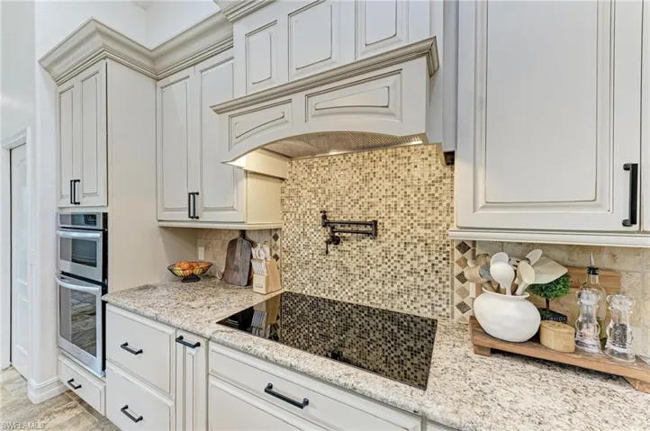 Kitchen with black electric cooktop, tasteful backsplash, light stone countertops, stainless steel double oven, and white cabinets